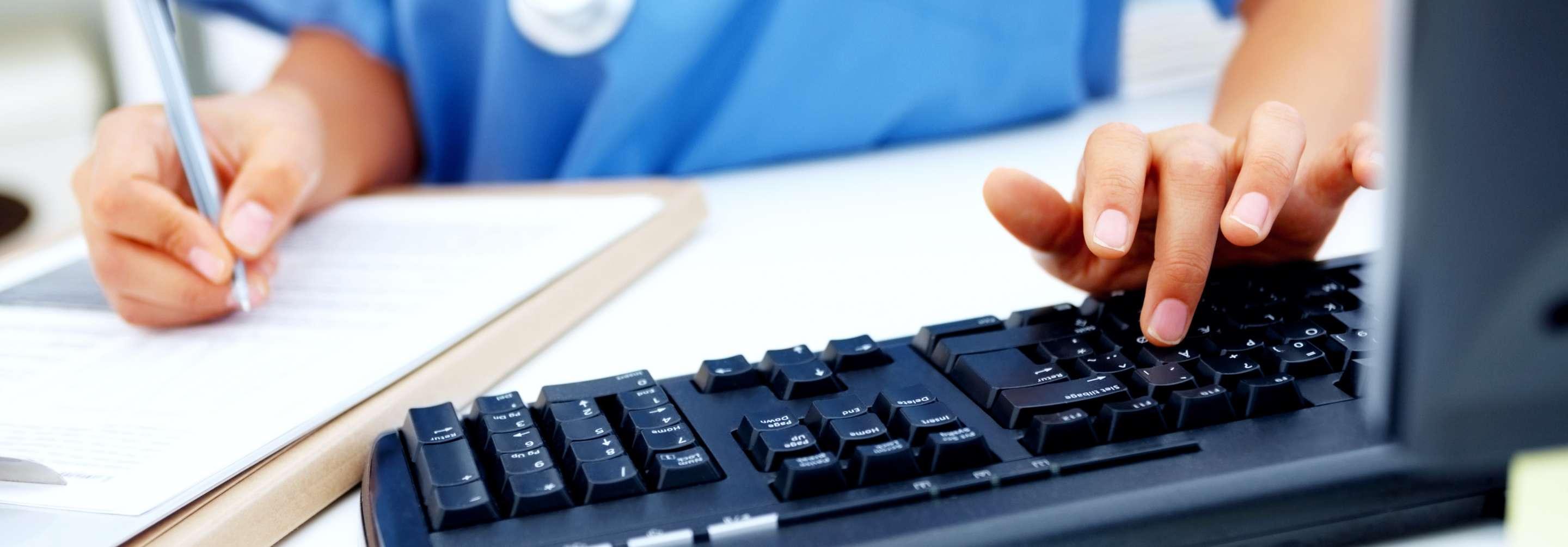 A person in blue scrubs works at a desk, writing notes on paper as well as typing on a keyboard