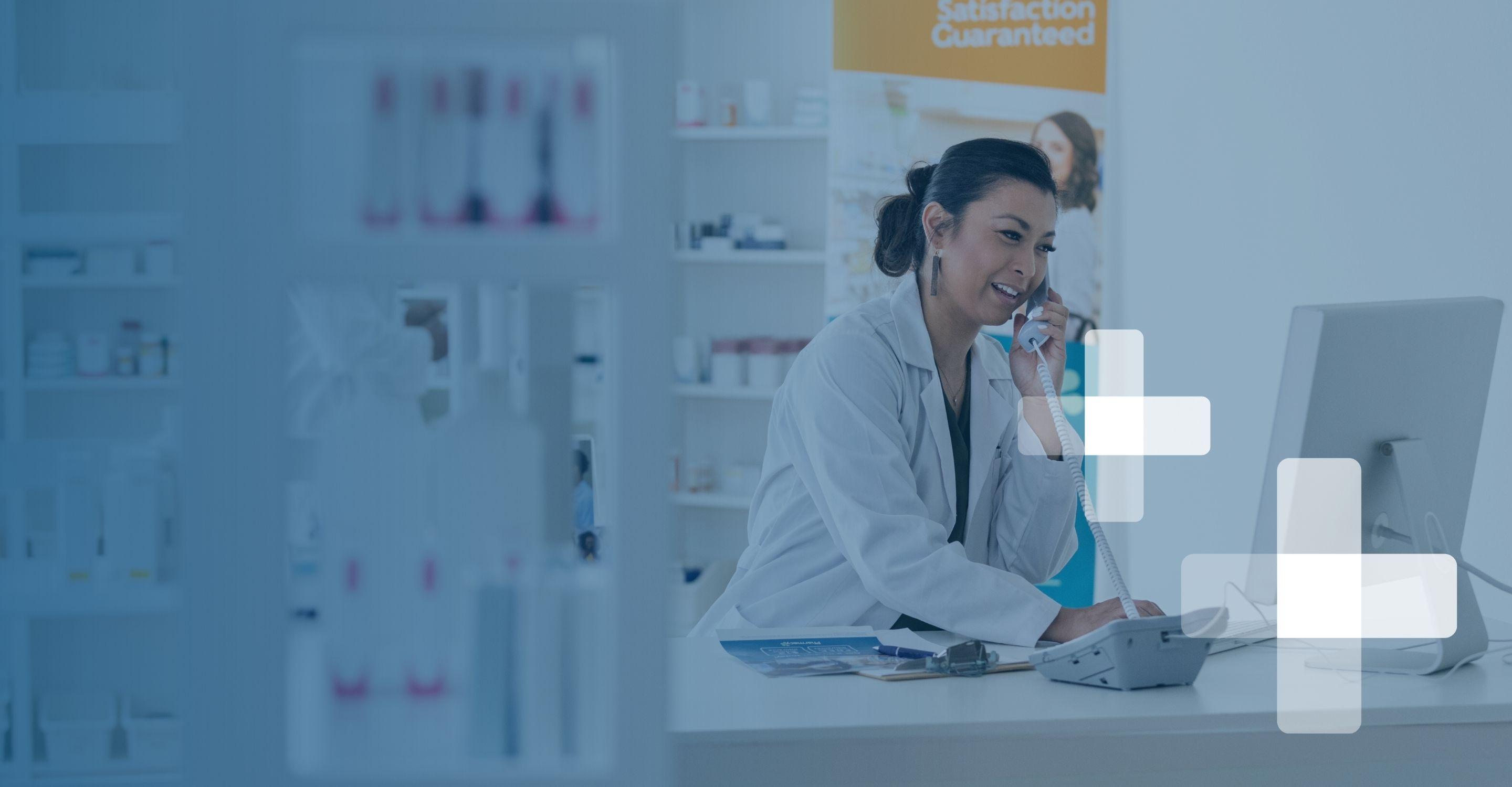 A female pharmacist in a white coat answers the phone at the front desk with a smile
