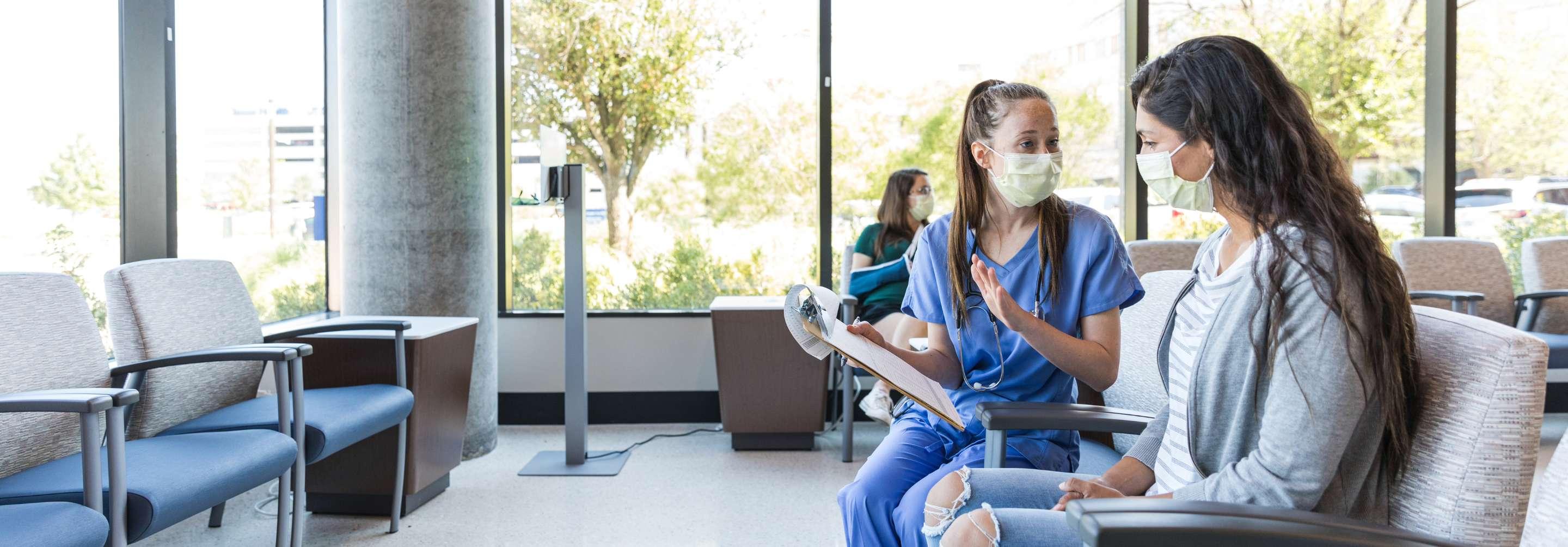 Triage nurse in blue scrubs and a surgical mask speaking with a patient in the waiting area