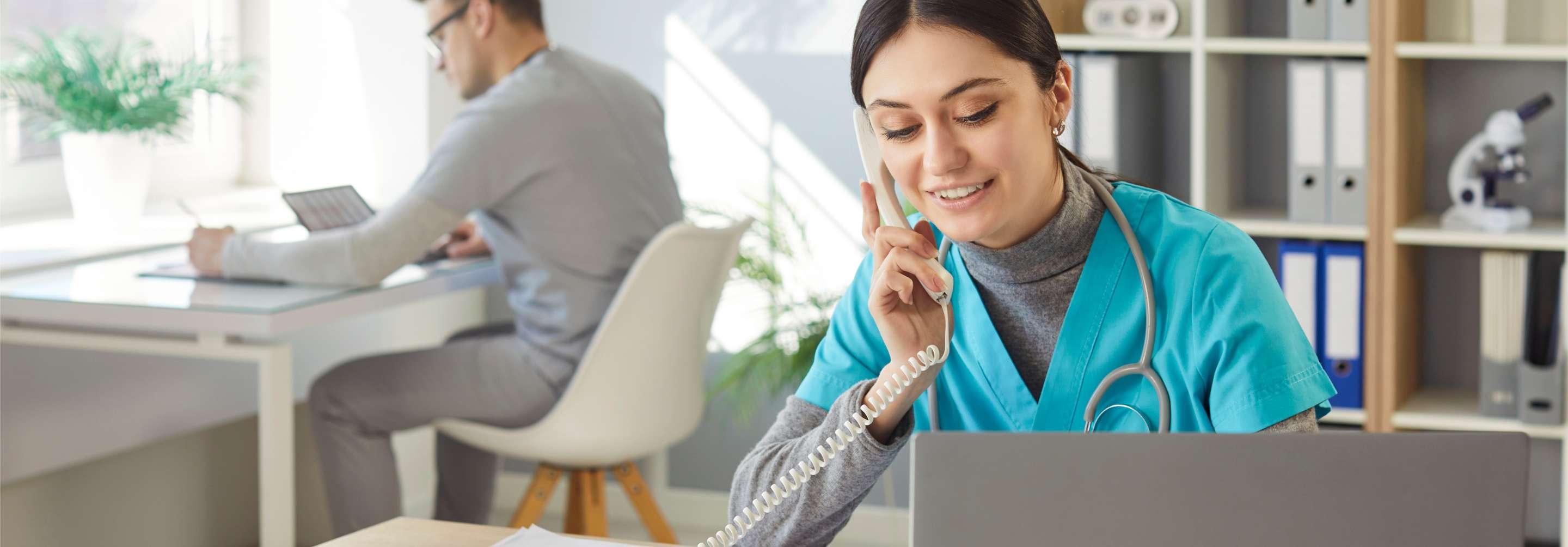 Utilization management nurse in blue scrubs speaking with someone on the phone while looking at the computer screen