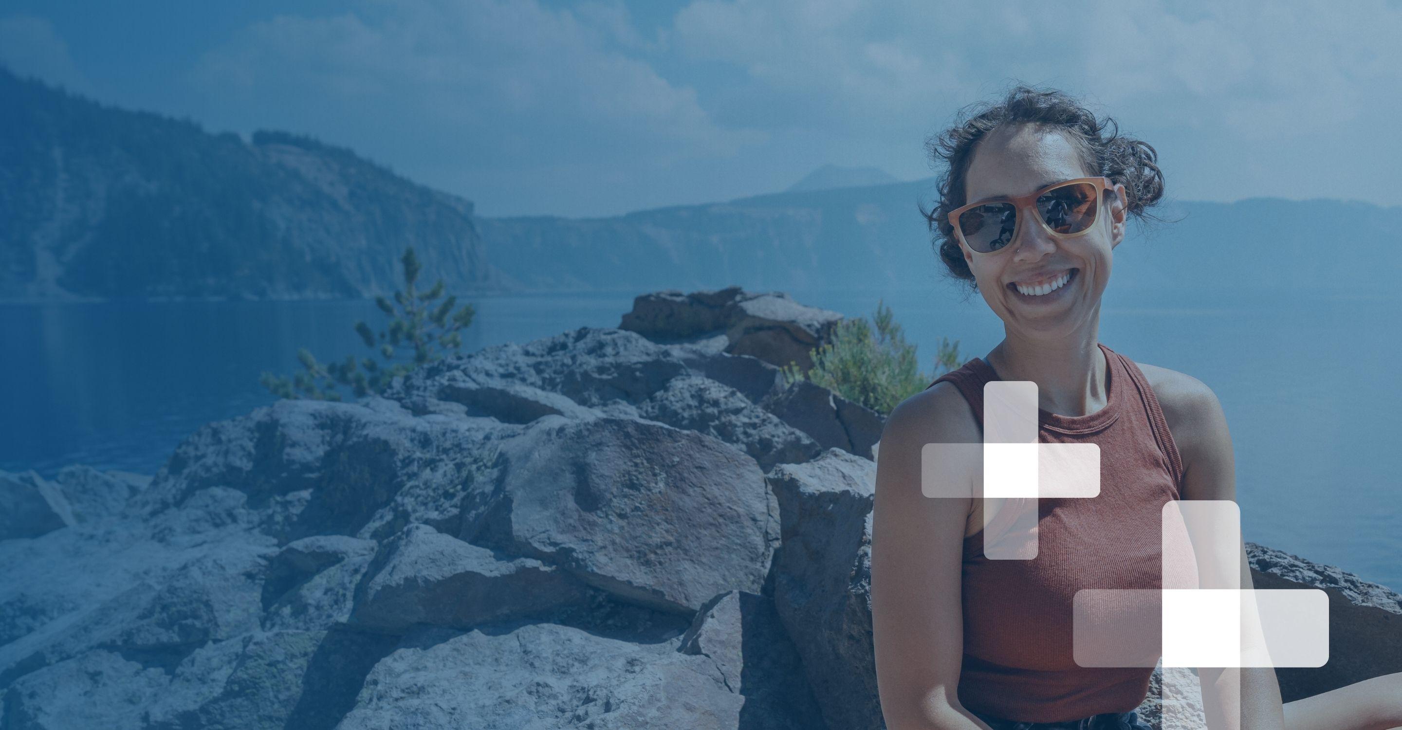 Person stands by a lake with rocky hills and forest in a scenic outdoor setting.