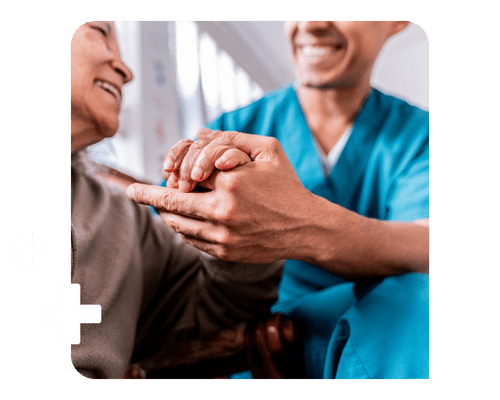 A healthcare professional in blue scrubs leans forward to give his hand to a smiling patient to assist them with mobility