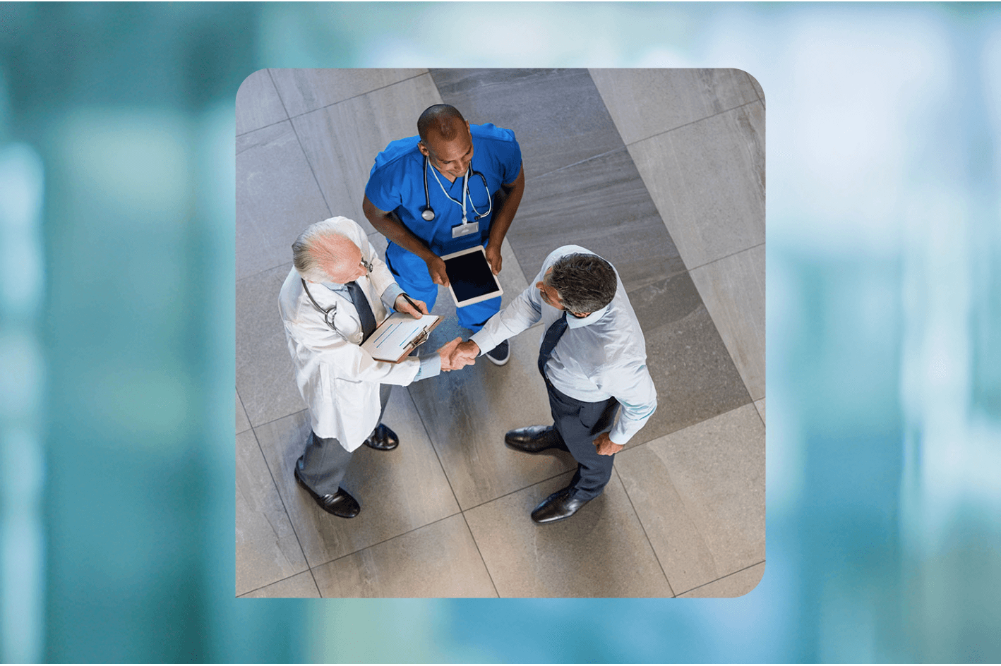 Three men meet in a facility lobby - a physician in a white coat shakes the hand of a man wearing a shirt and tie, while a man in scrubs looks on with a tablet