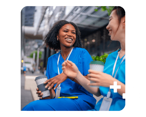Two female healthcare professionals in scrubs sit on a bench laughing and drinking coffee