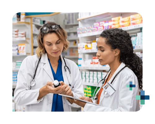 Two pharmacists in lab coats discuss medication while reviewing notes in a pharmacy aisle.