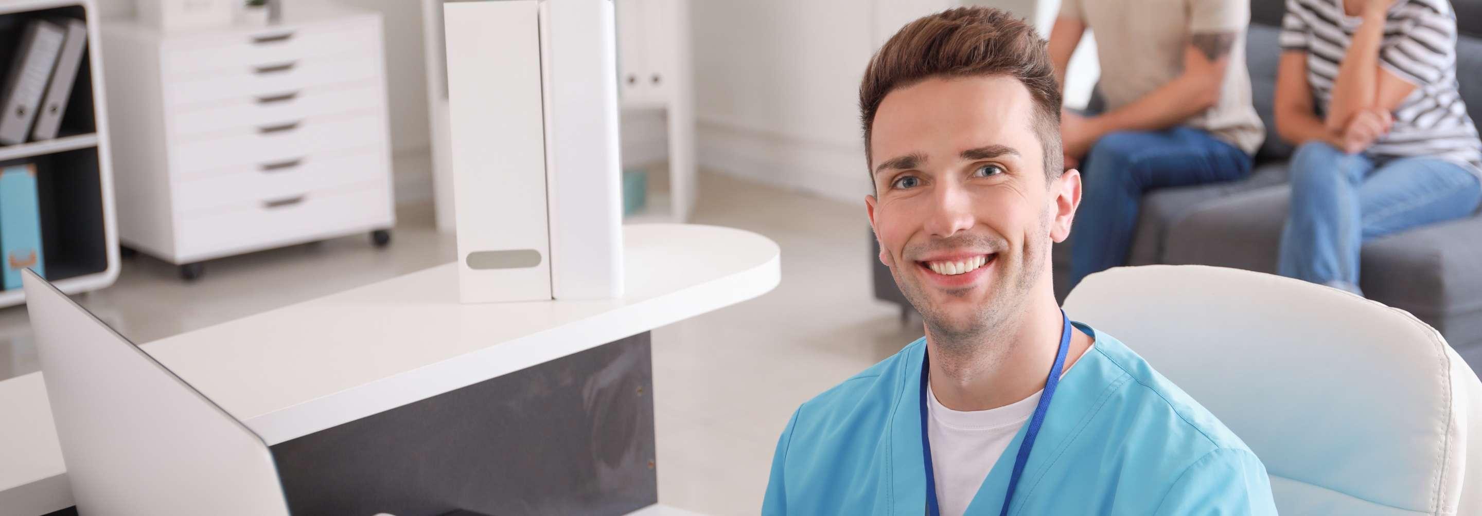 Patient access representative in blue scrubs smiling at the camera, sitting at a computer in an office area with patients in the background