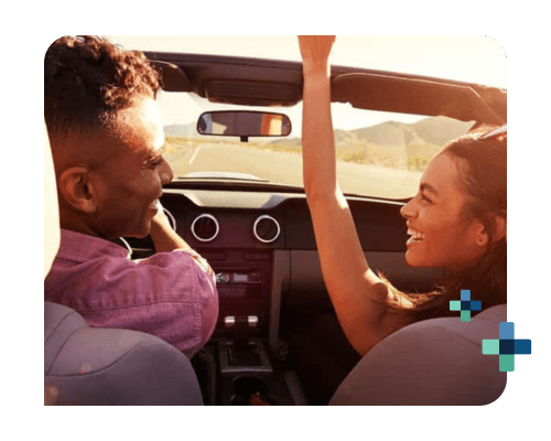 Two people in a convertible on a sunny road trip with mountains in the background.
