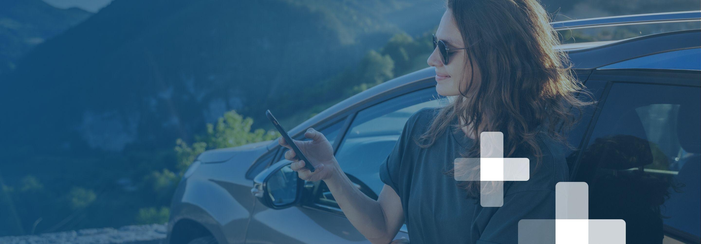 A young woman in sunglasses stands outside the drivers' side of her sedan and checks her cellphone in front of mountain scenery