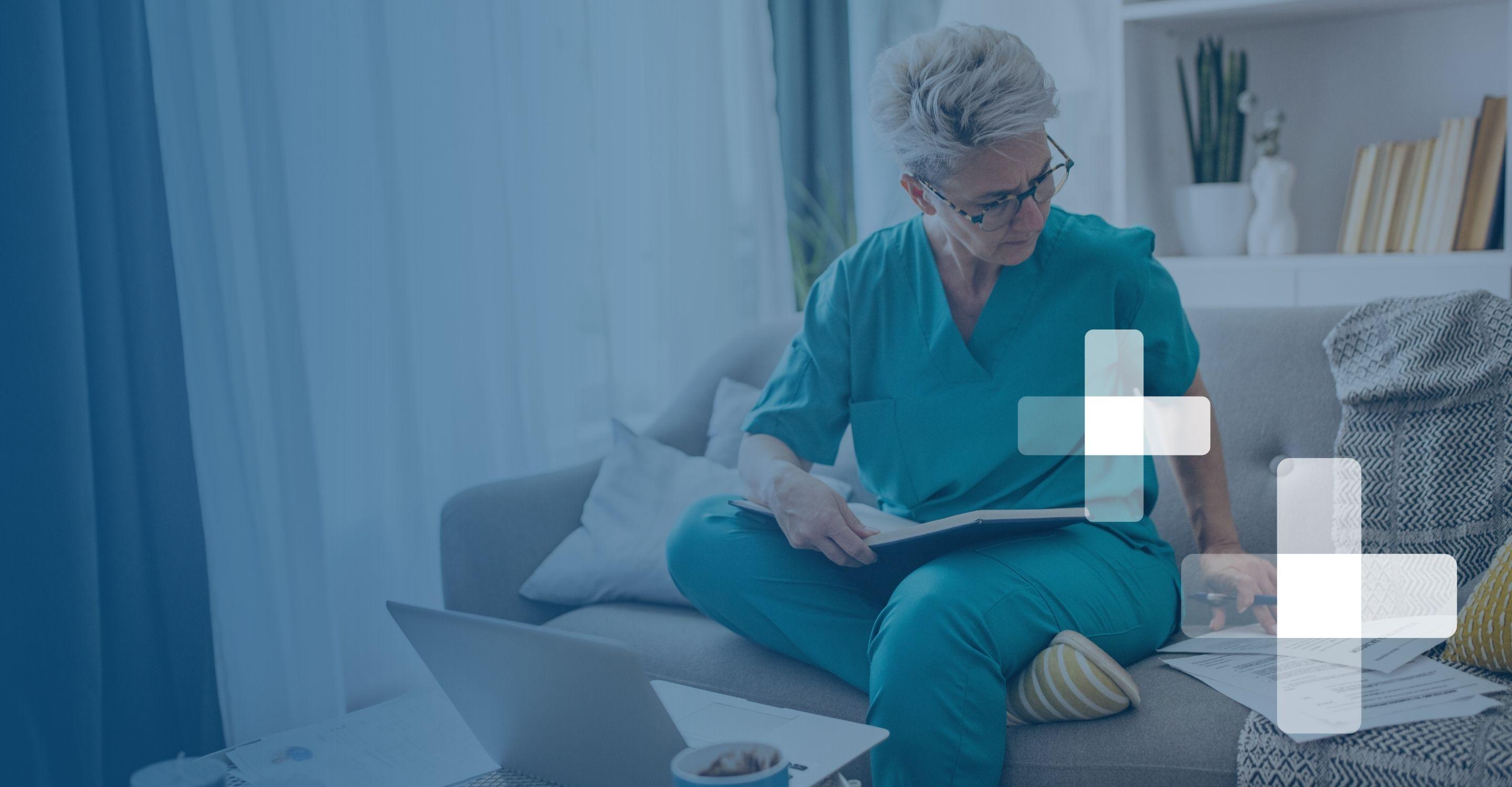 An older nurse in blue scrubs and glasses studies books and papers on her couch in front of a laptop
