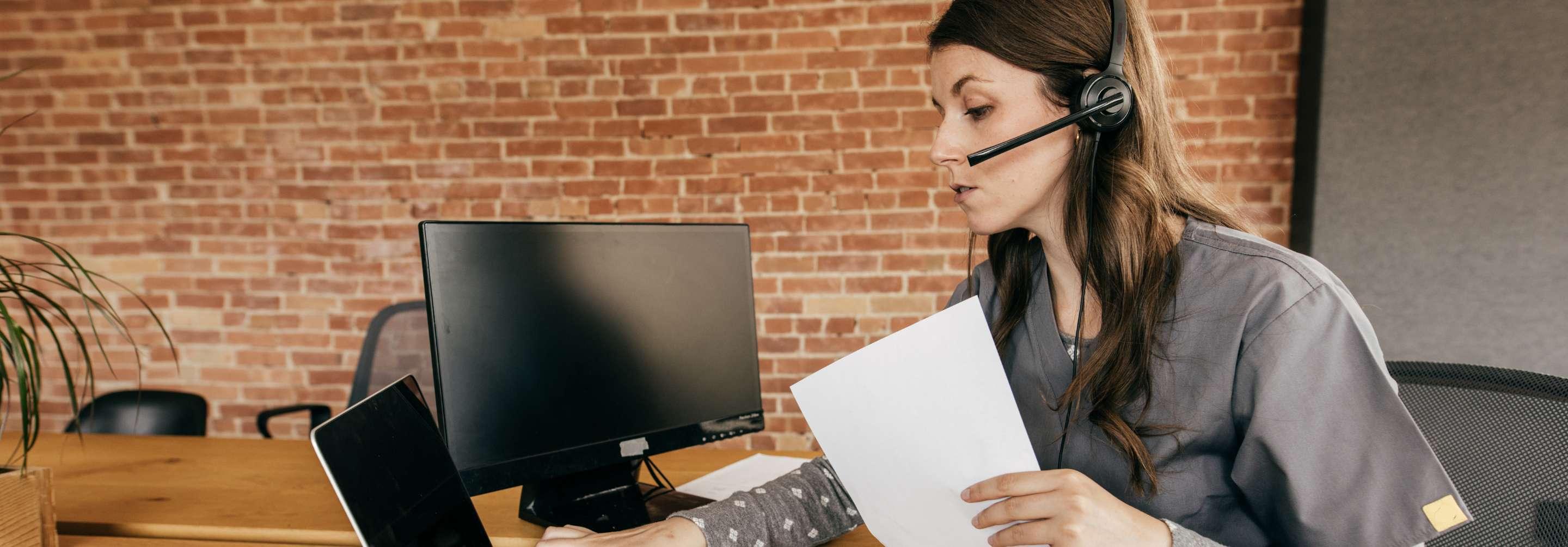 Prior authorization nurse in grey scrubs processing papers, wearing a headset and working at the computer