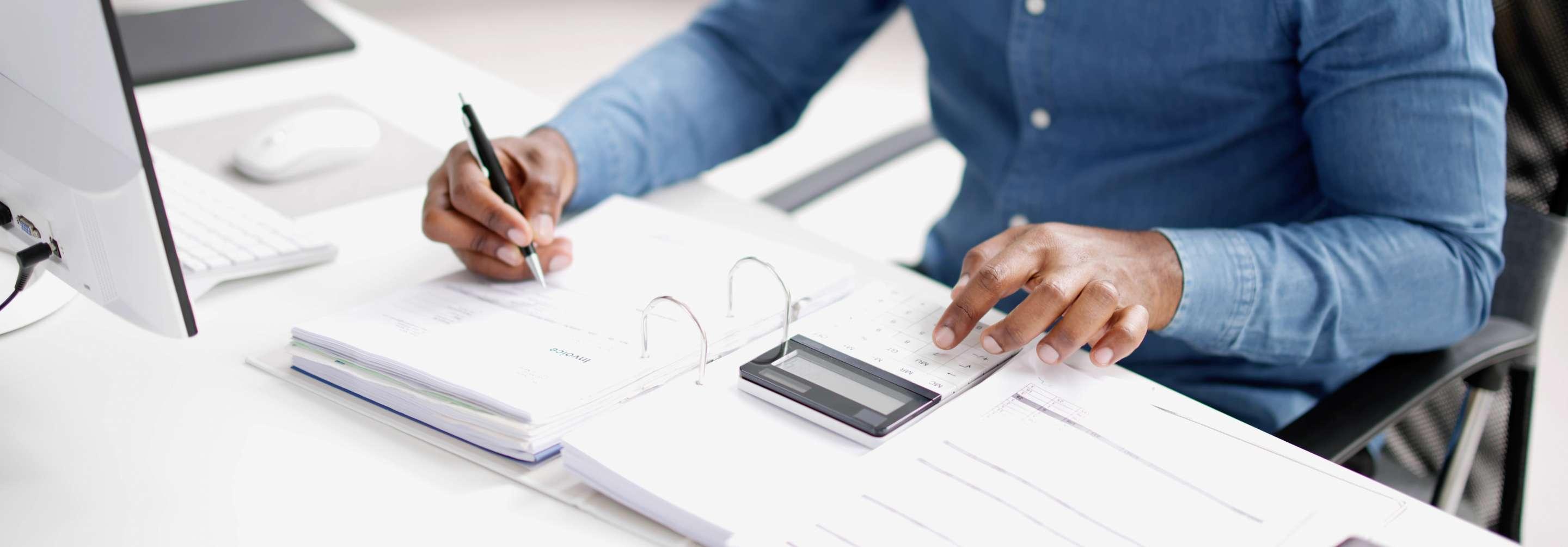 A healthcare accounting clerk sitting at a desk tabulates numbers on a calculator